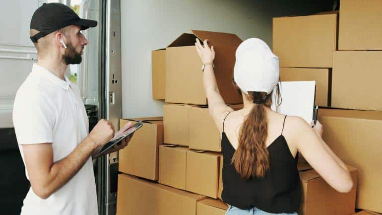 Woman stacking cardboard boxes in a moving truck as a mover holds a clipboard and tracks inventory.
