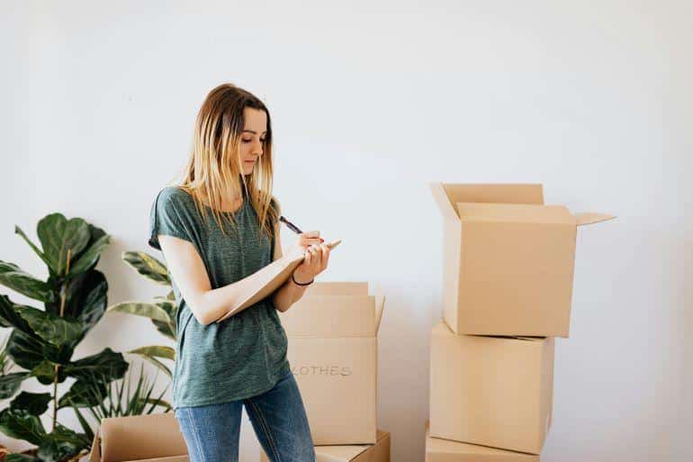 Woman writing notes on a clipboard beside stacked cardboard boxes during packing and organization.