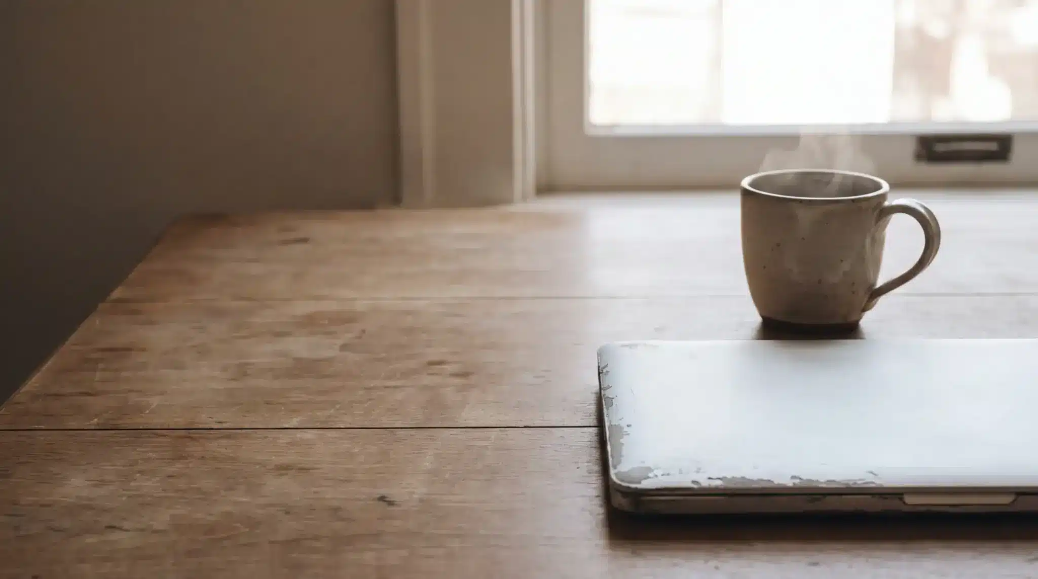 Steaming mug next to closed laptop on rustic wooden table in warm lighting