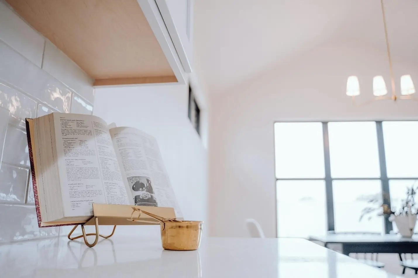 Open cookbook on a stand in bright kitchen with gold measuring cup nearby