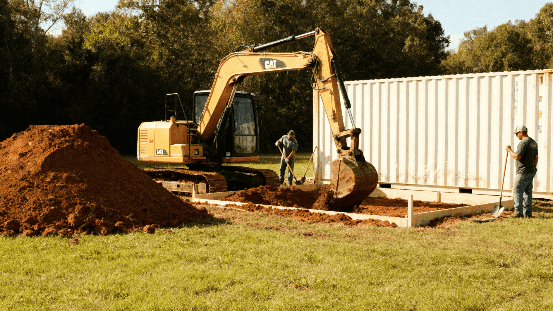 excavator digging foundation beside shipping container while workers shovel dirt and prepare base for construction site
