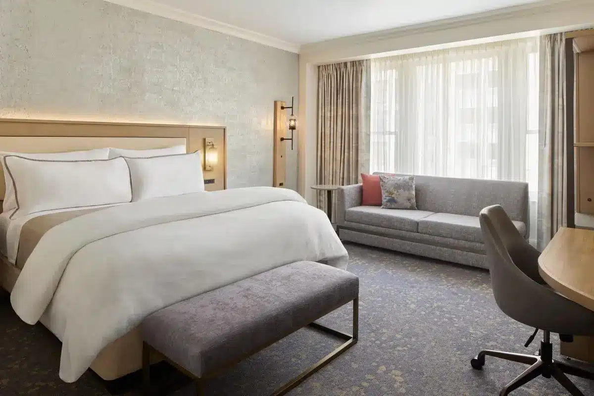 Modern hotel room with king-size bed, grey sofa, and desk under natural light