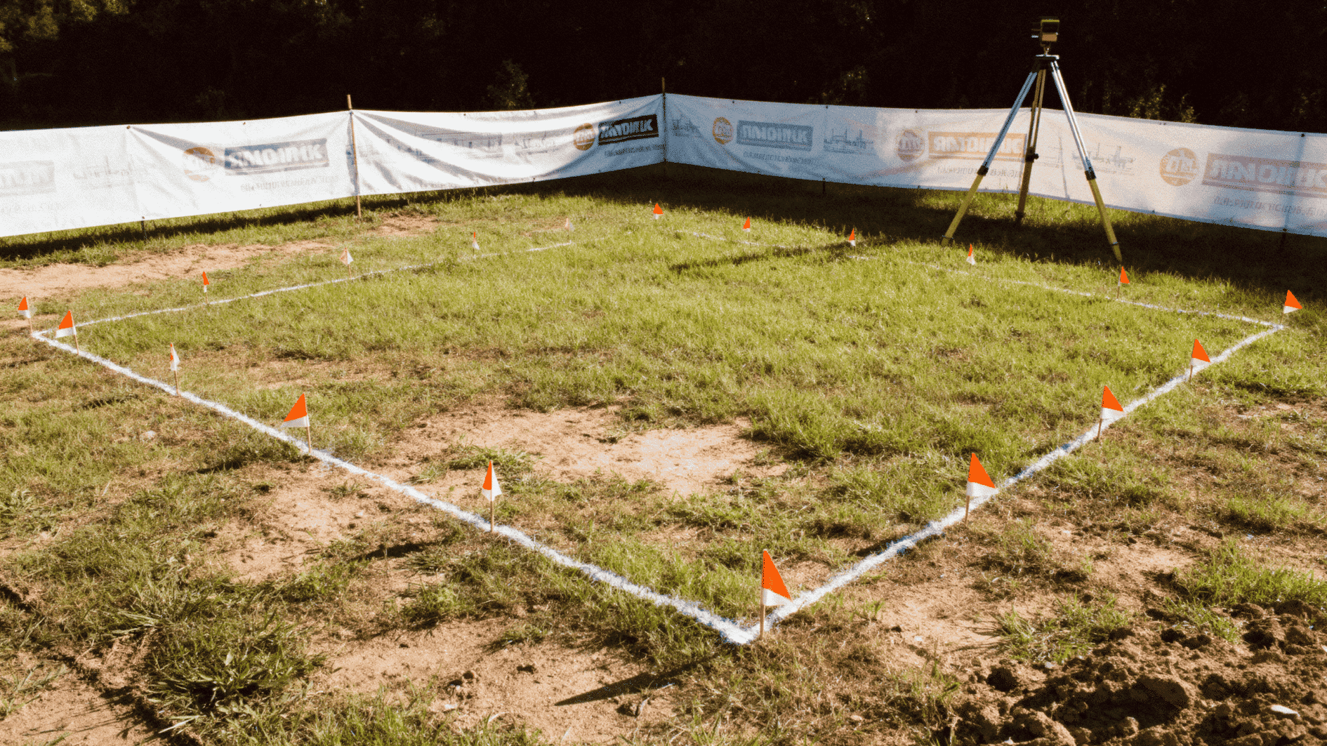 grassy land marked with white lines and small orange flags forming rectangular boundary, surrounded by safety banners