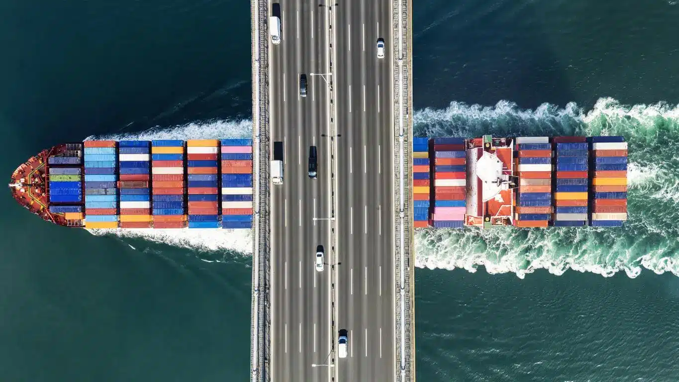 Cargo ship carrying multicolored containers passing under a busy highway bridge