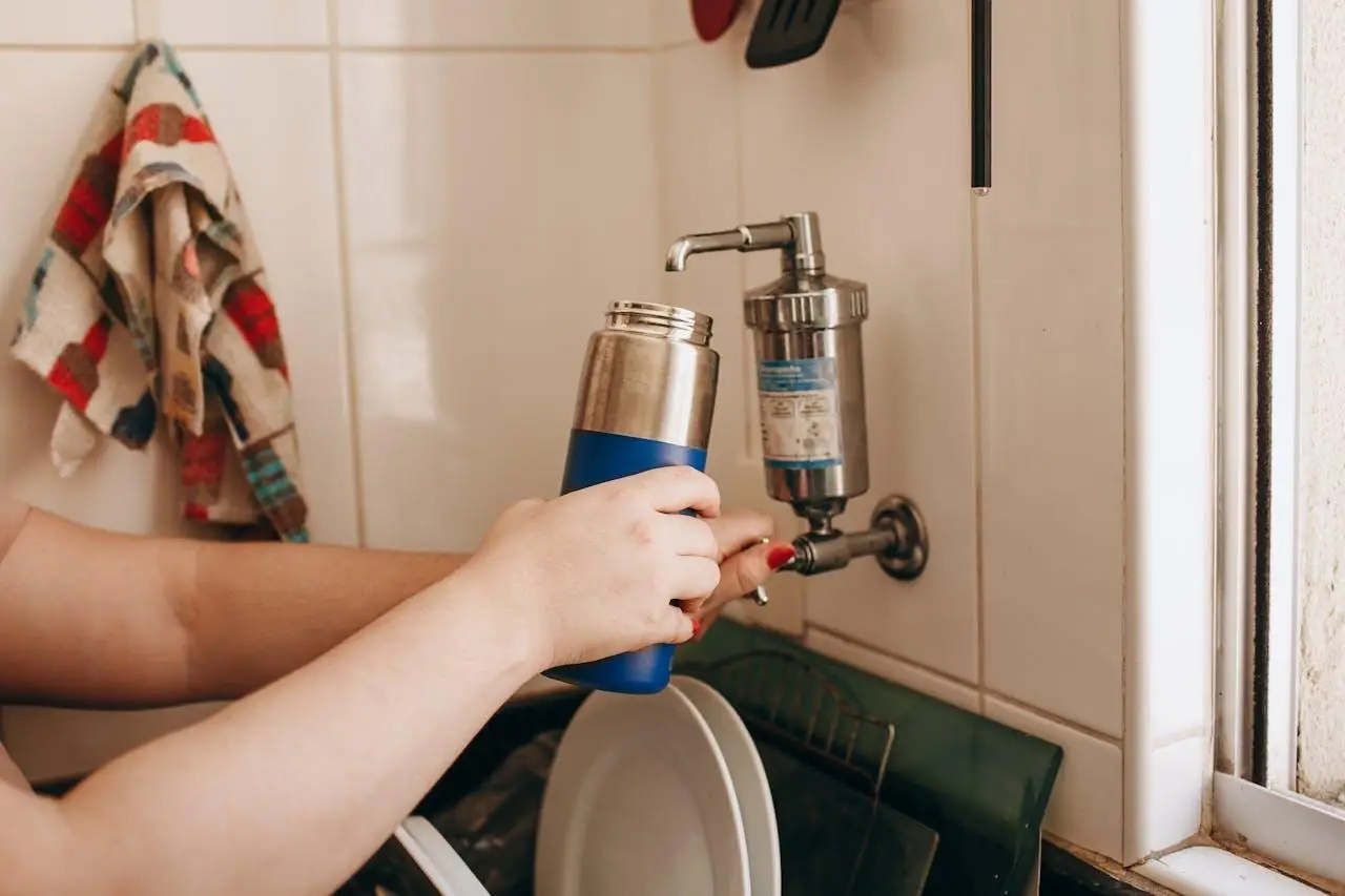 Person filling blue thermos from kitchen water filter near dish rack
