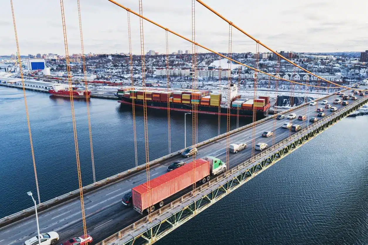 Cars and truck crossing suspension bridge with container ship and city in background