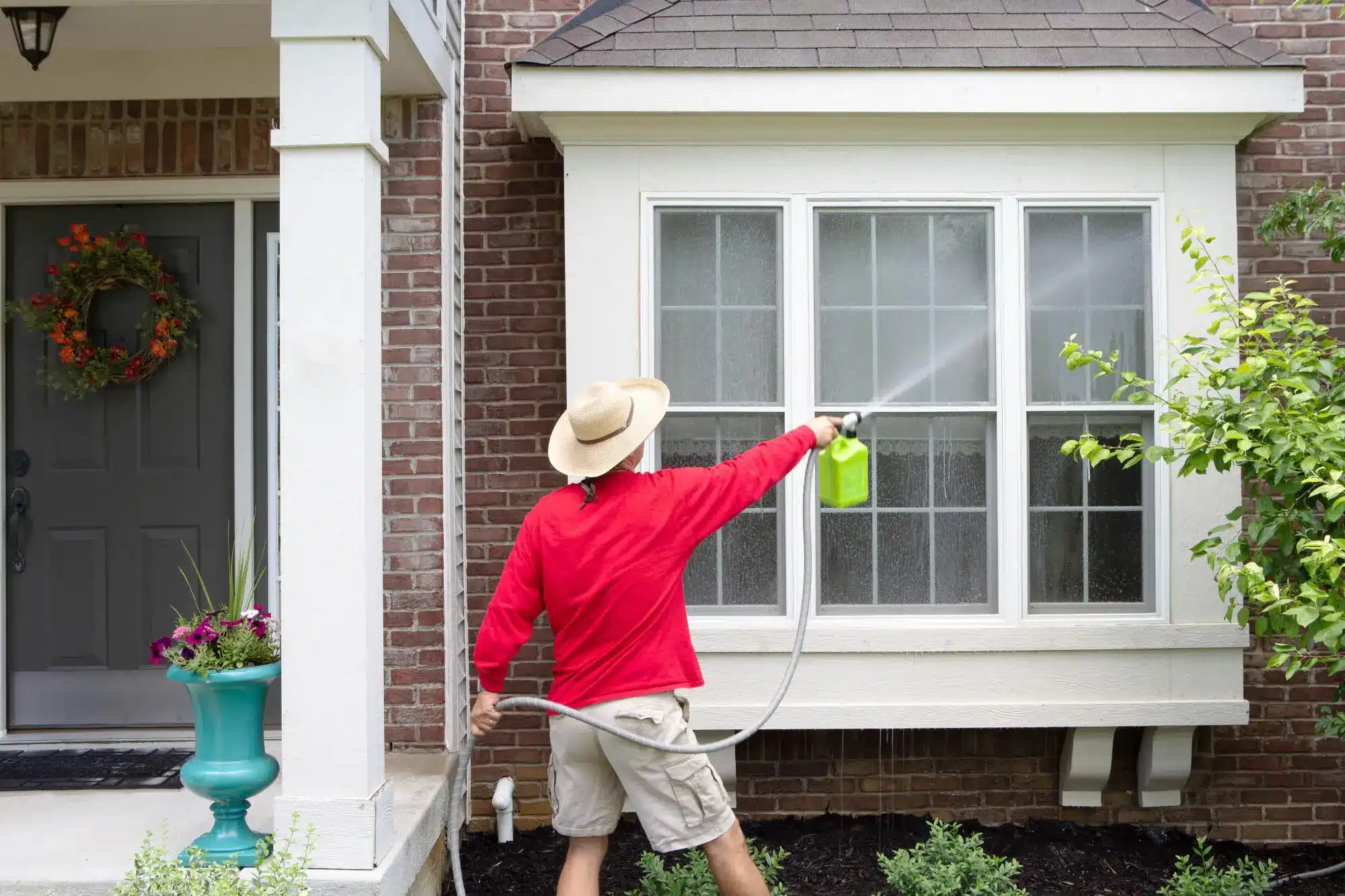 Man in red shirt cleaning house windows with garden hose in brick exterior setting