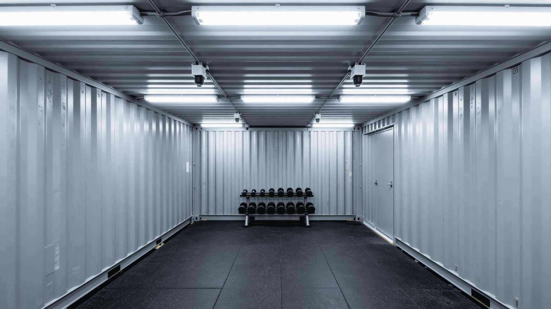 minimalist shipping container gym interior with rubber flooring, overhead lights, and a dumbbell rack centered against wall