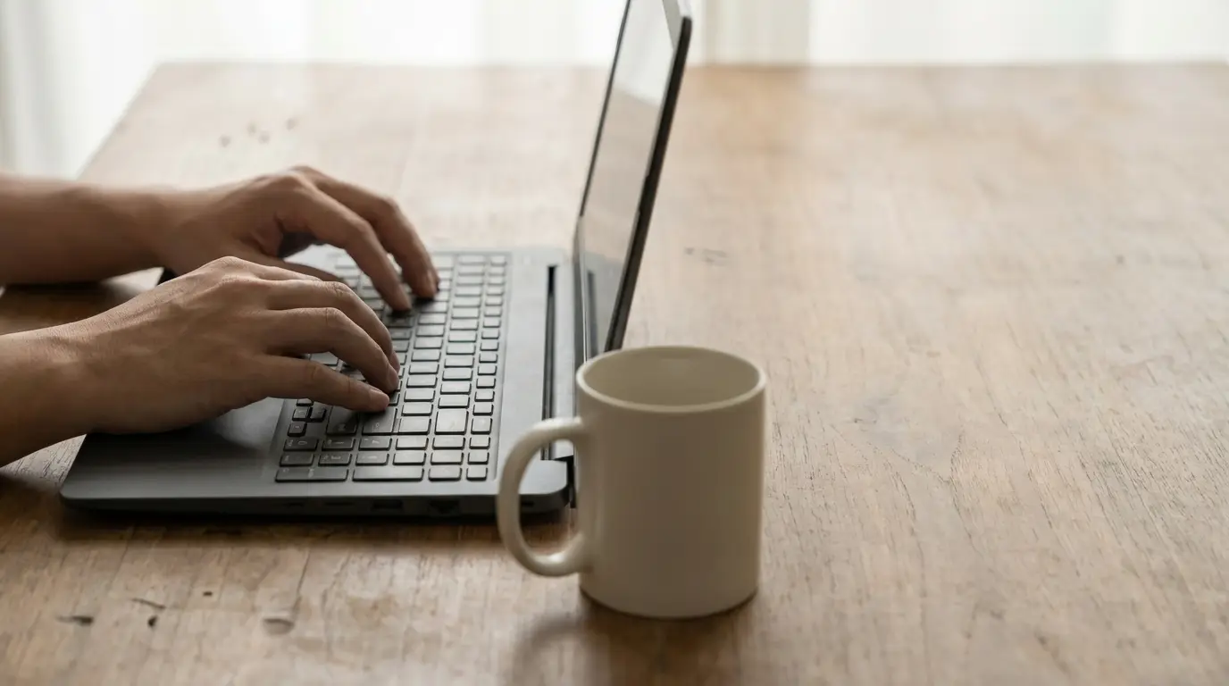 Hands typing on a laptop keyboard with a white mug on a wooden table