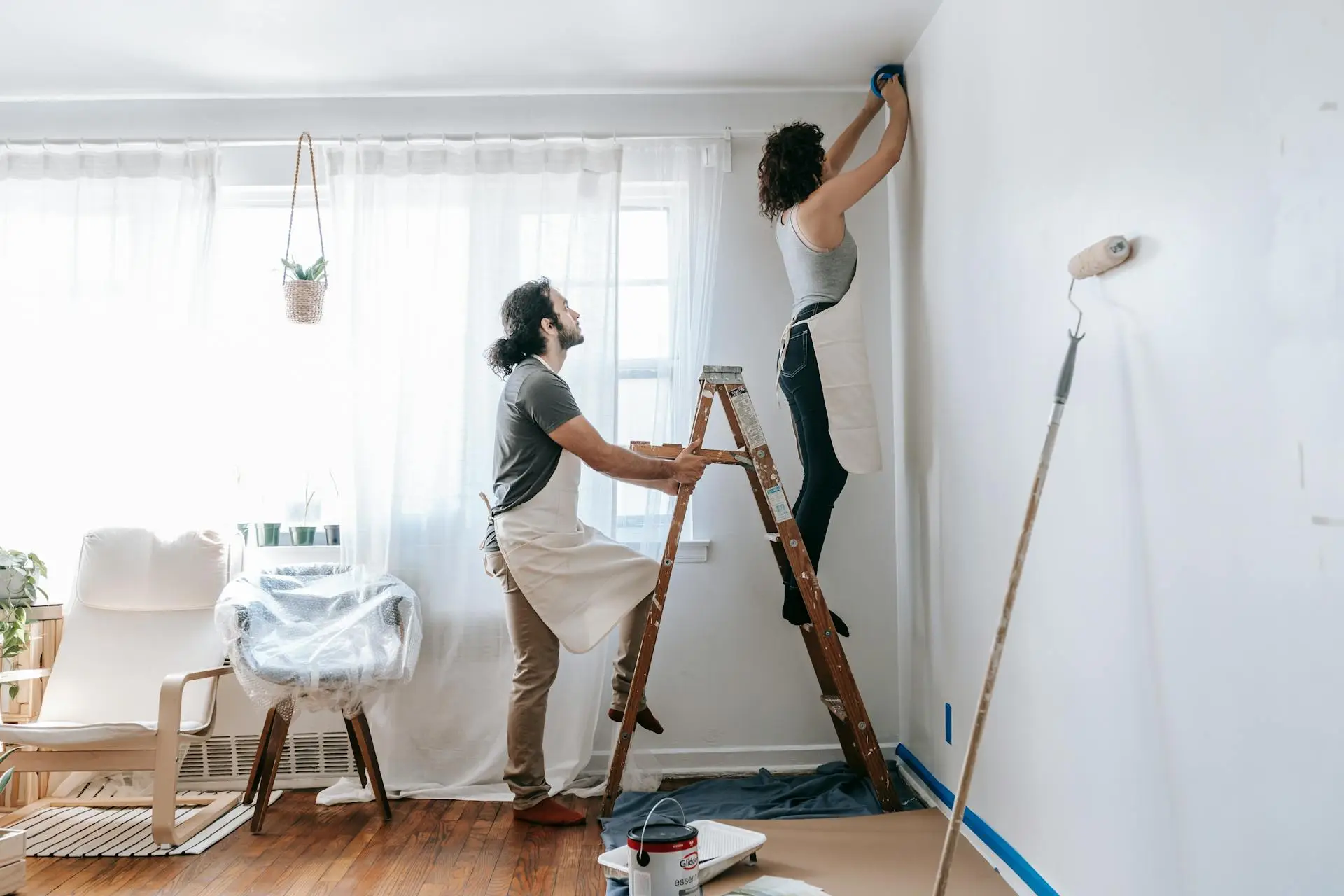 Two people wearing aprons painting a white wall with ladder in bright living room