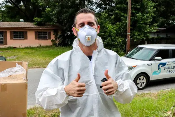 Person in protective coveralls and mask giving thumbs up outdoors near parked van