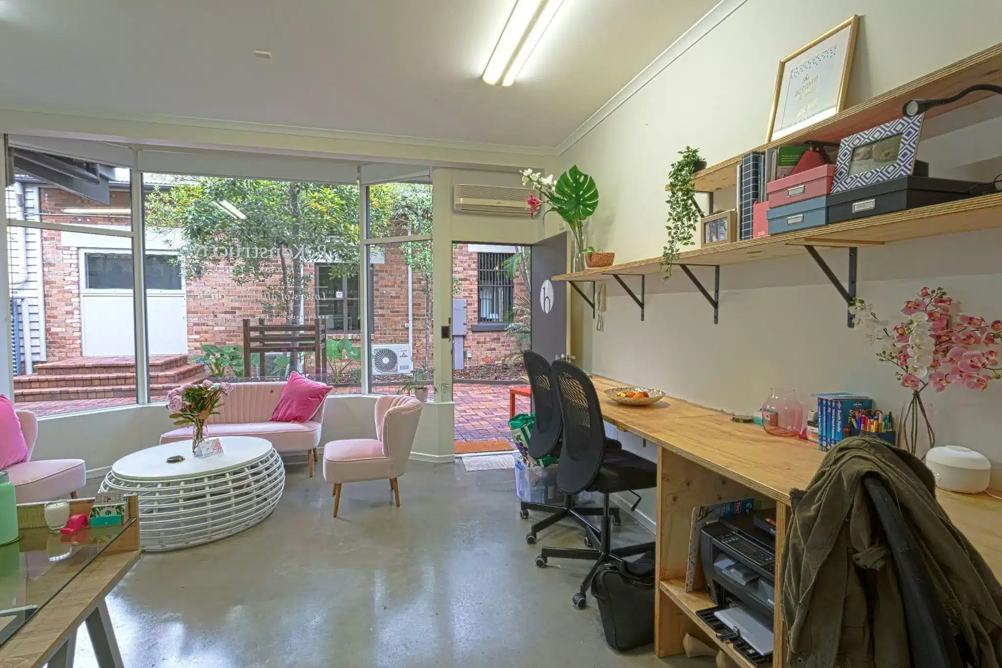 Modern office with pink chairs, wooden desk, and wall shelving, overlooking a brick courtyard