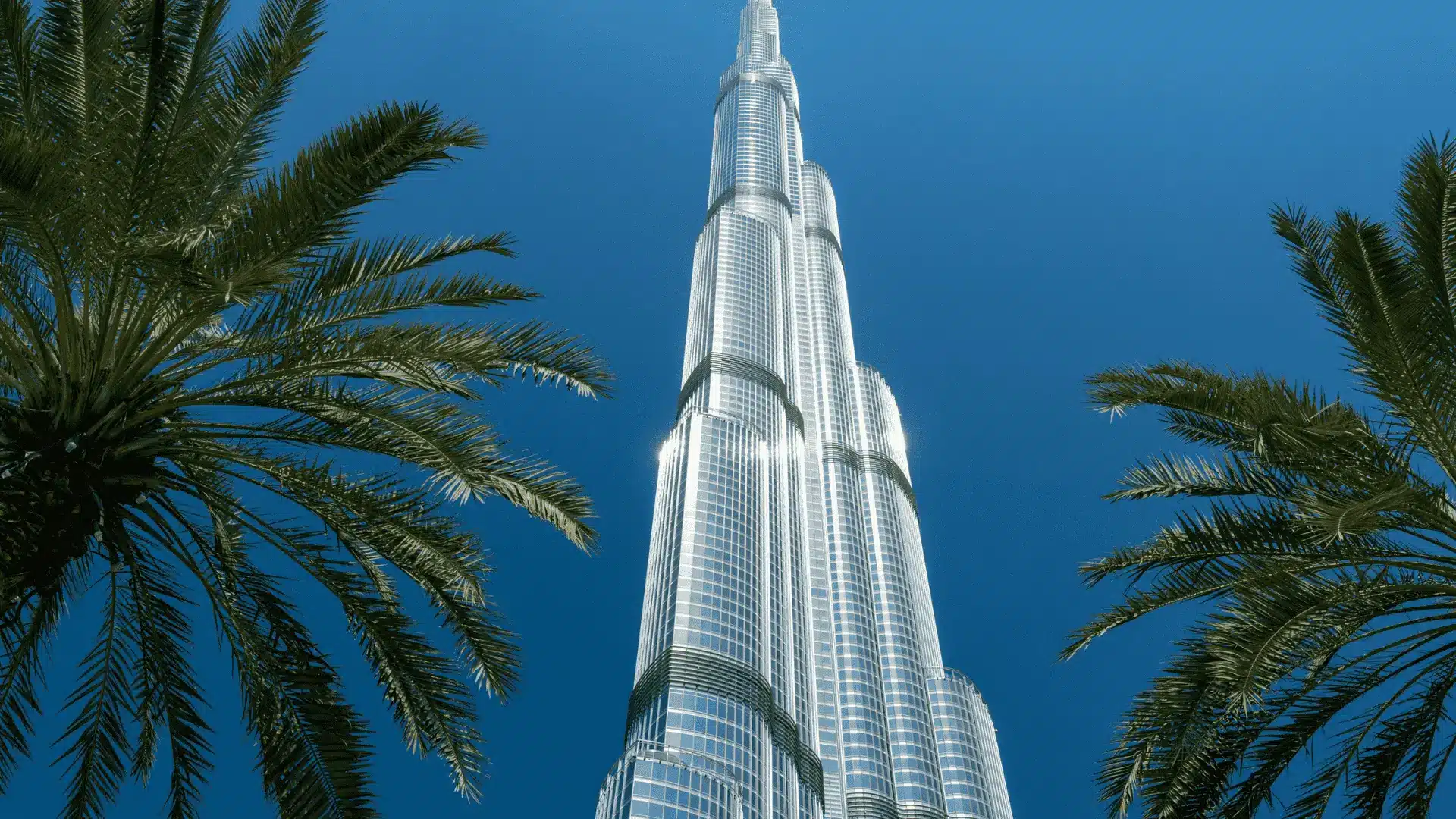 Burj Khalifa in Dubai, towering above palm trees with its sleek glass exterior against a clear blue sky.