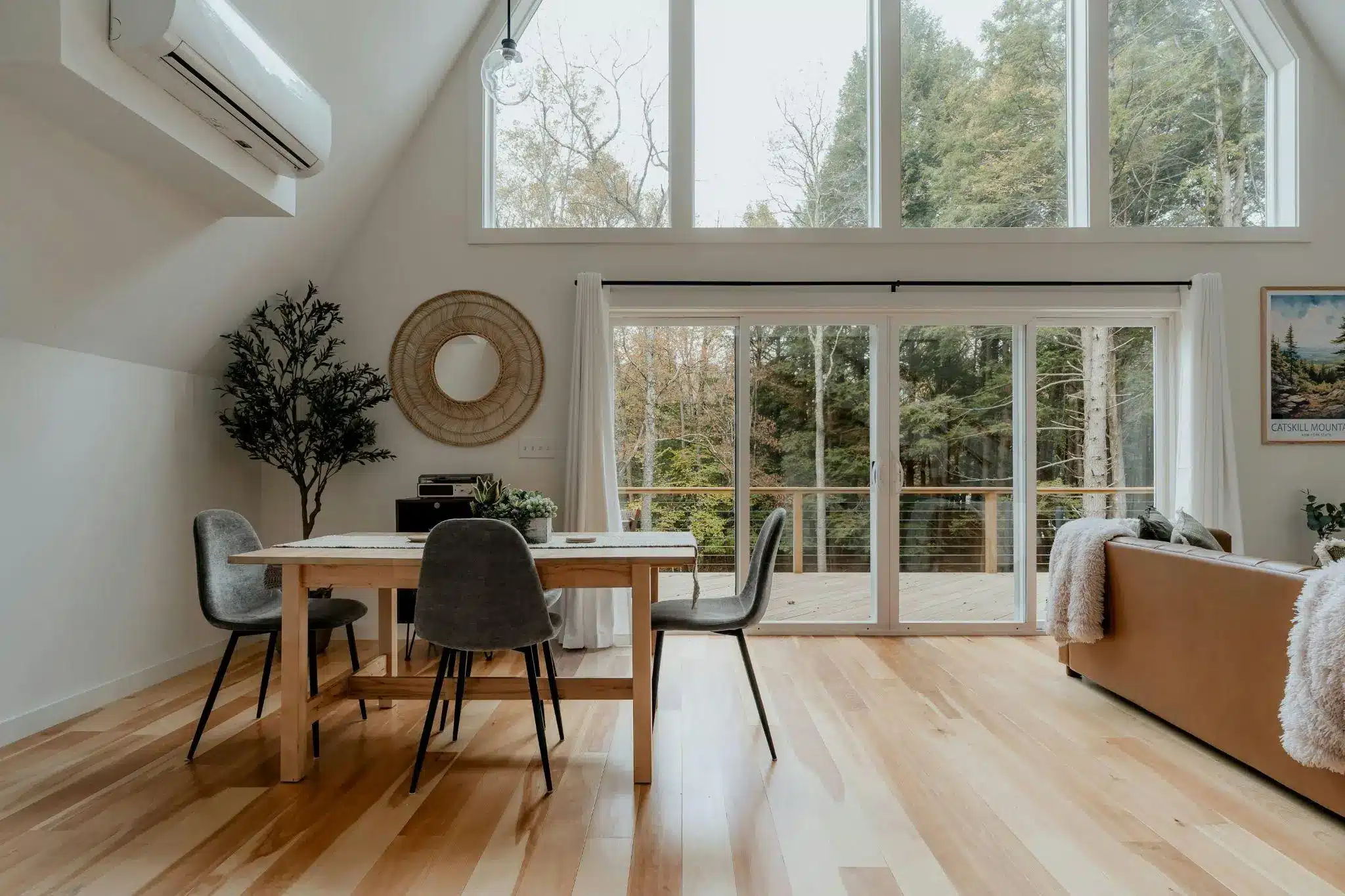Open-plan living space with wooden dining table, gray chairs, and large glass doors overlooking forest