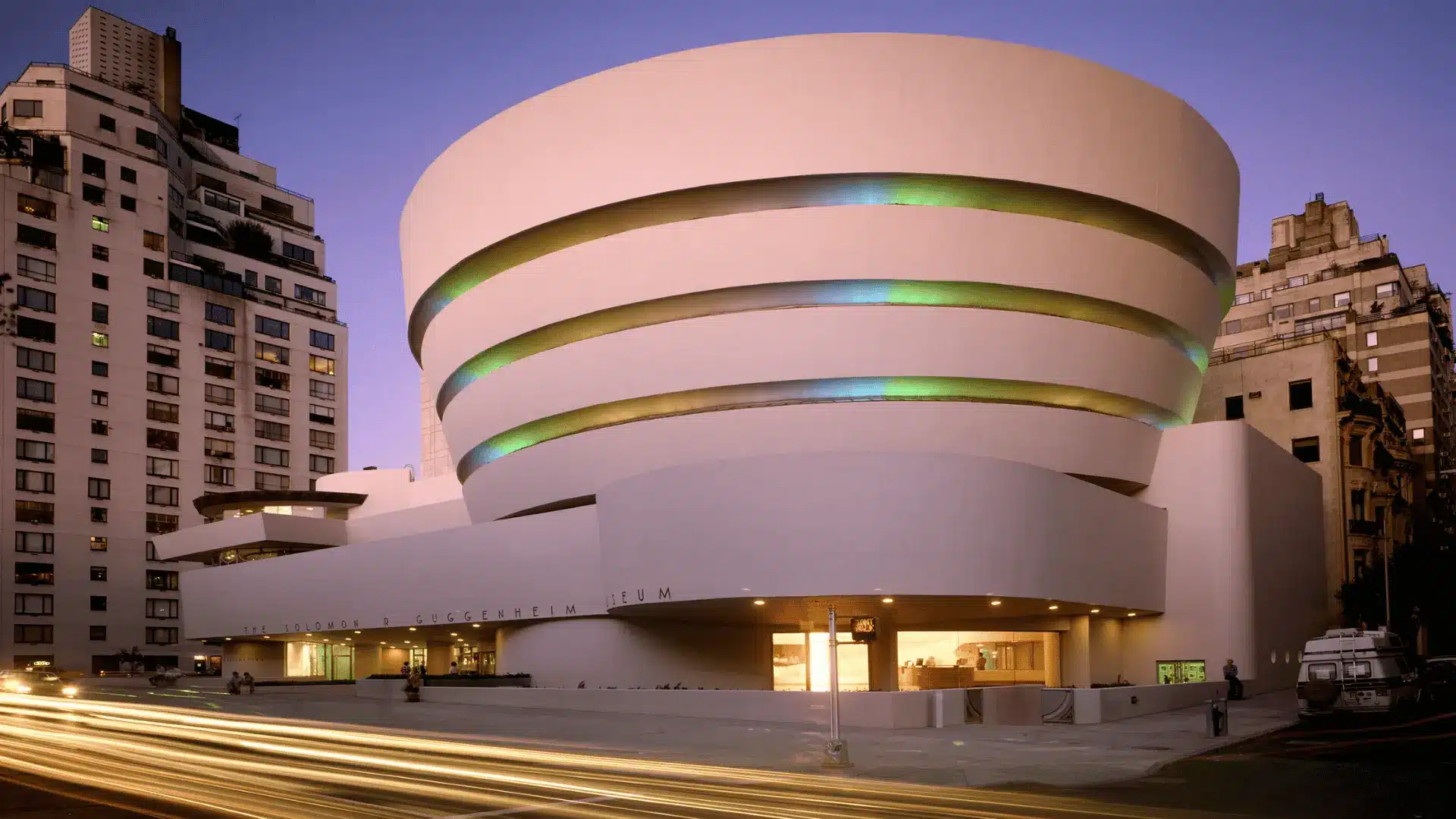 Guggenheim Museum in New York with a spiraling, smooth white exterior, set against a cityscape at dusk with lights and traffic.