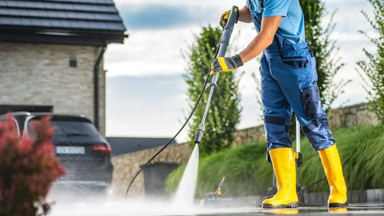 Person pressure washing driveway outdoors wearing yellow boots and blue work attire