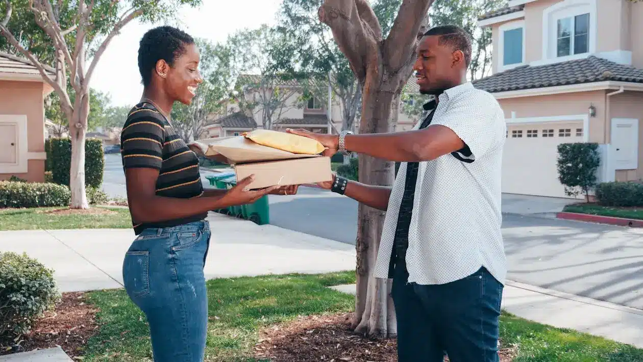 Two people exchanging parcel boxes on a suburban sidewalk, surrounded by trees and houses