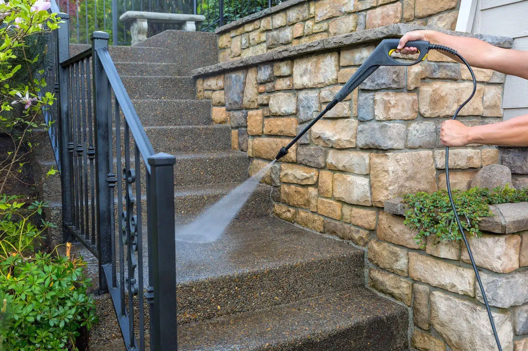 Person pressure washing stone steps with black metal railing and stone wall in outdoor setting