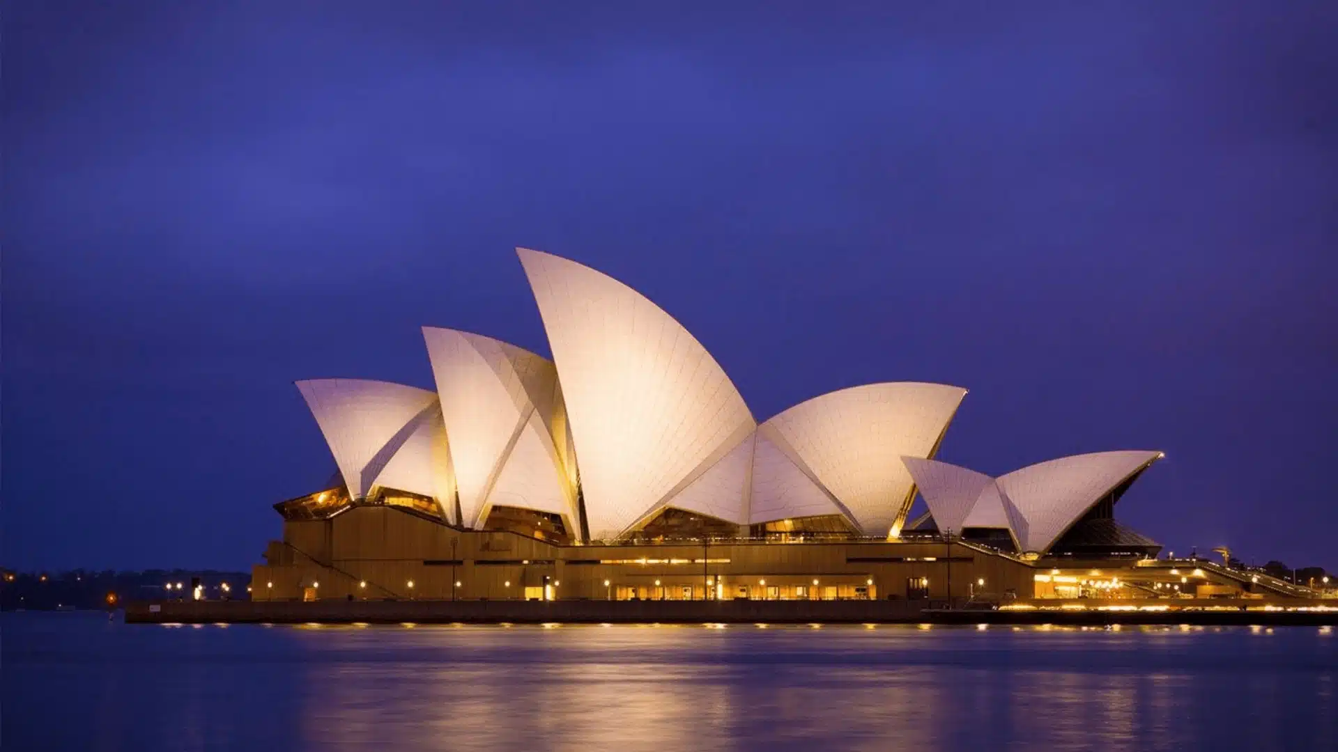 Sydney Opera House at night, with its iconic sail-like design illuminated against the dark sky, reflecting in the water.
