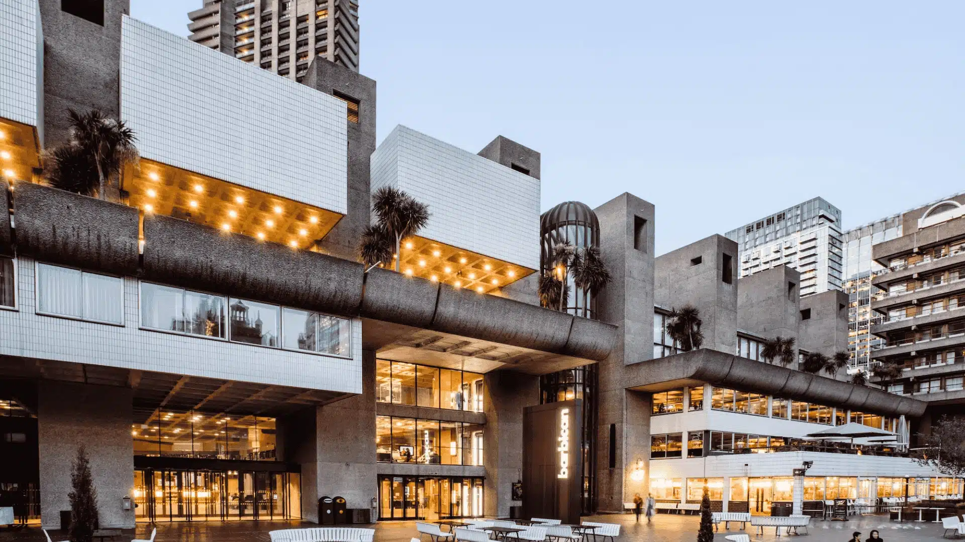 The Barbican Centre in London, showcasing Brutalist architecture with bold concrete forms, illuminated edges, and a bustling public plaza.
