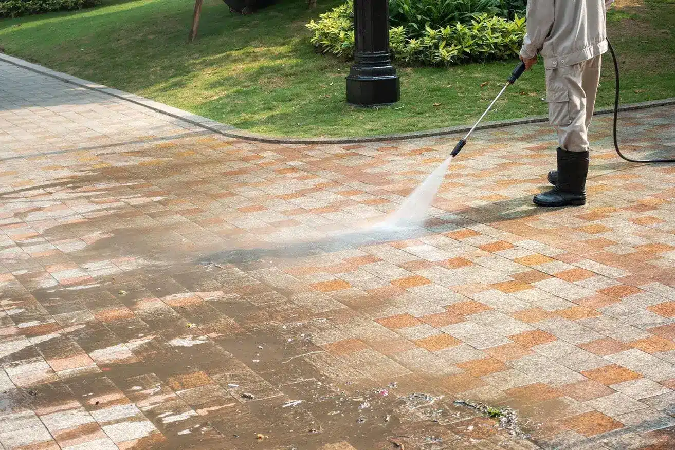 Person pressure washing a tiled outdoor pathway with nearby green lawn and plants
