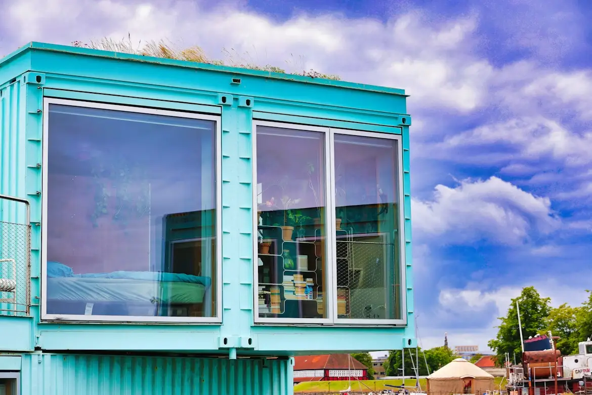 Turquoise shipping container home with large windows and rooftop plants under blue sky