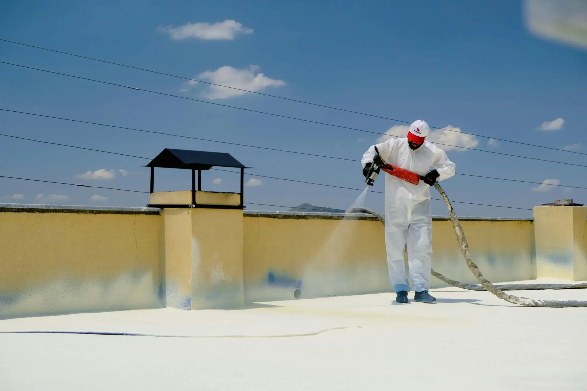 Worker in protective suit spraying foam insulation on rooftop under clear blue sky