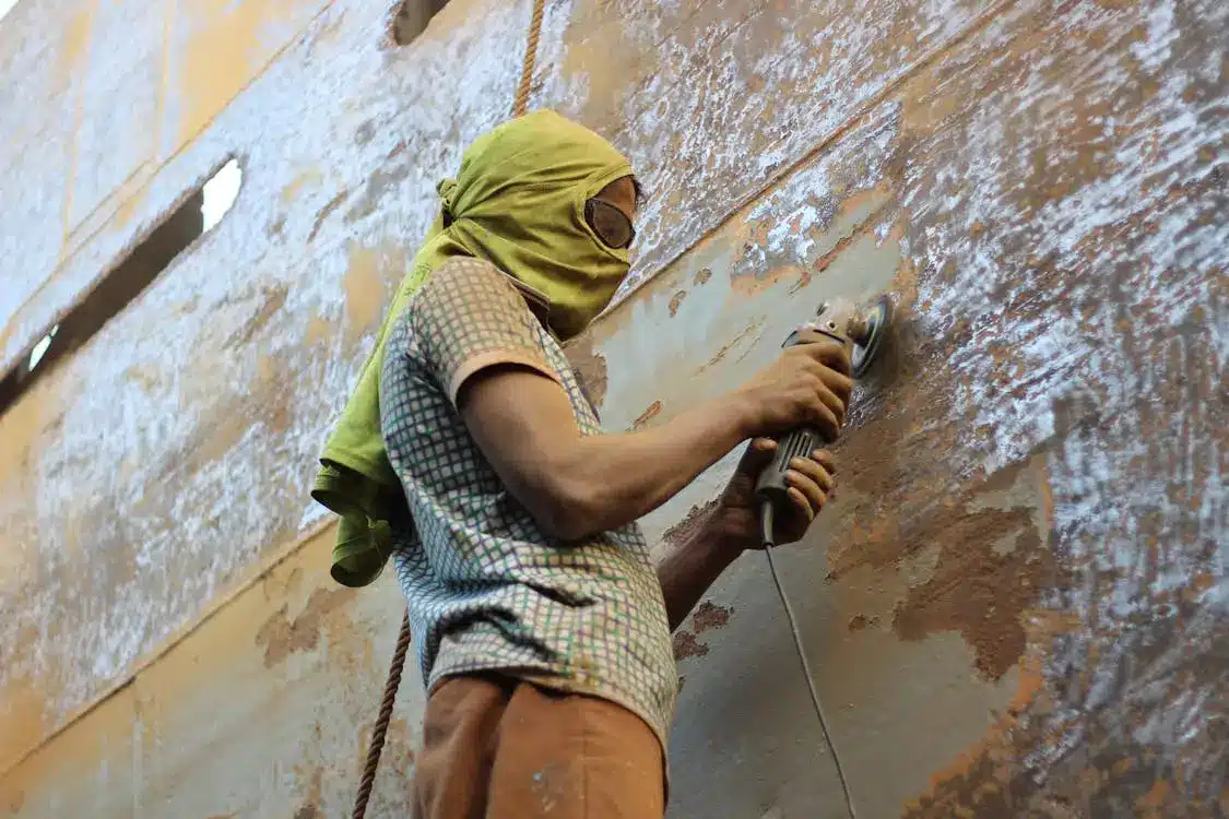 Worker wearing headscarf and goggles using power tool on rusty metal surface outdoors