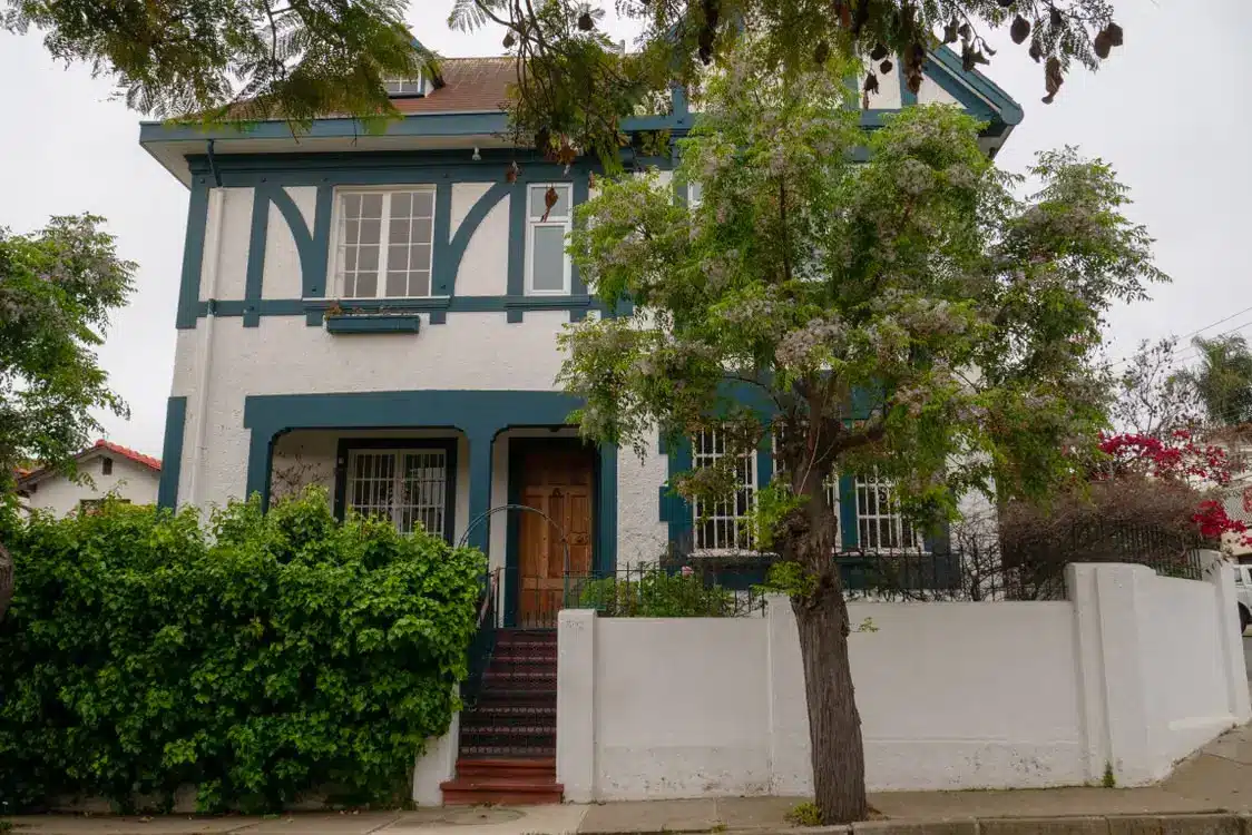 Two-story house with blue trim and wooden door surrounded by greenery