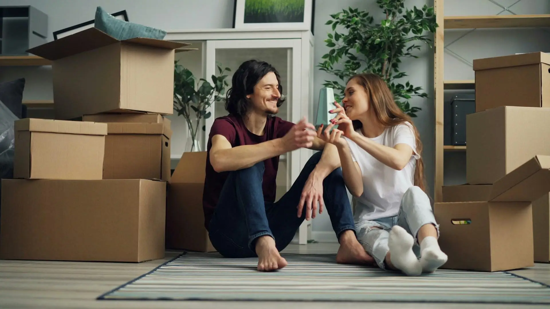 Couple sitting on rug surrounded by cardboard boxes in living room with plants and shelves