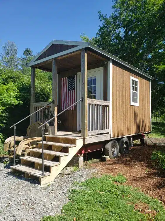 Tiny house on wheels with wooden porch and American flag in sunny garden setting
