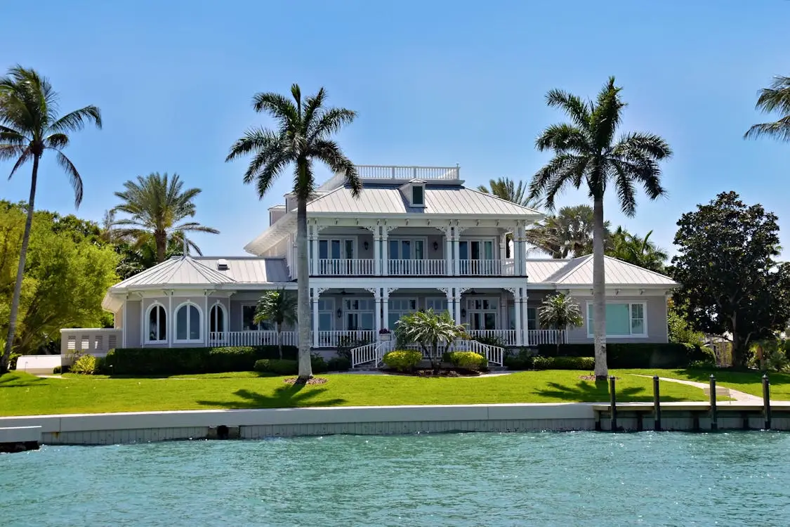 Colonial-style waterfront house with palm trees and manicured lawn under clear blue sky