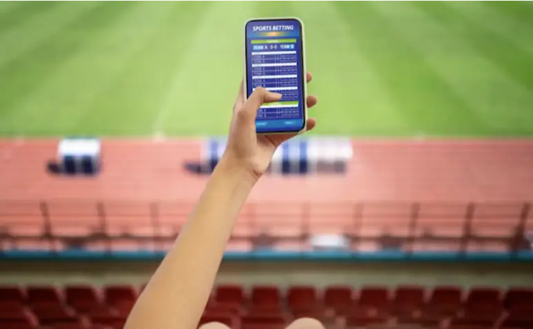 Hand holding smartphone displaying sports betting app in outdoor stadium with empty seats