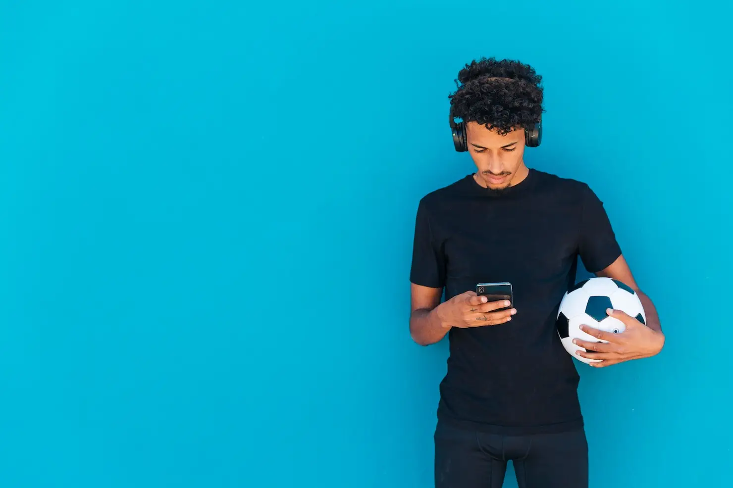 Man in black t-shirt with headphones holding soccer ball against blue wall