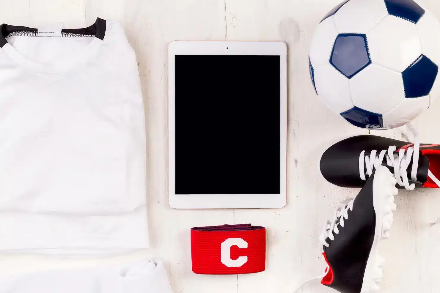 Soccer gear with uniform, cleats, tablet, and captain's armband on white wooden surface