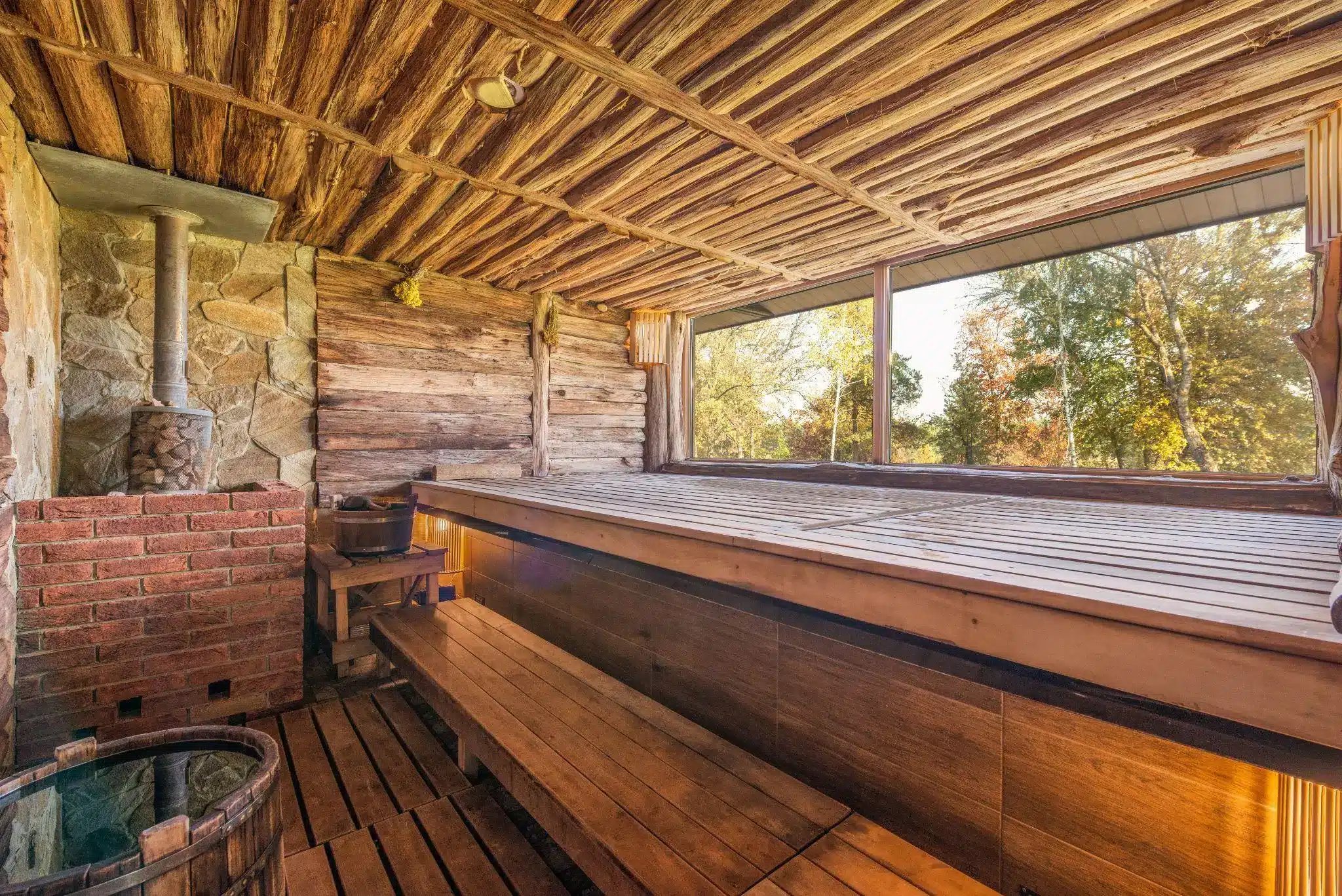 Rustic wood sauna interior with stone stove and large window overlooking trees