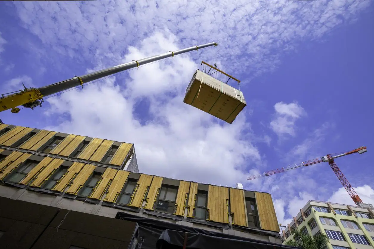 Crane lifting large crate against partly cloudy sky near construction site building