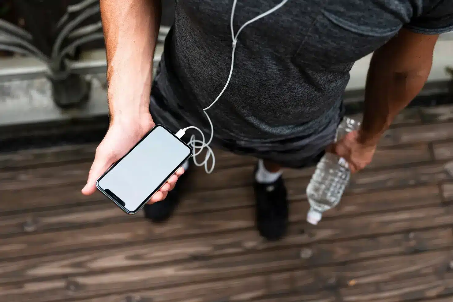 Person holding smartphone with earphones and water bottle on wooden deck