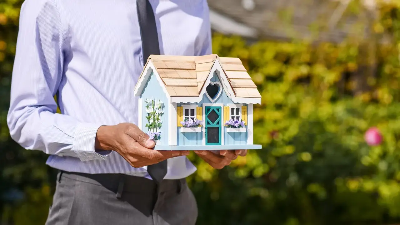 Person holding a miniature blue house with wooden roof in a sunny outdoor garden setting