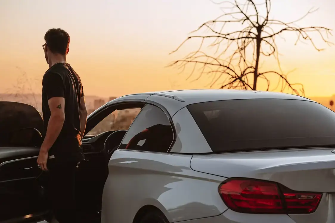 Man standing beside white car at sunset with bare tree in the background
