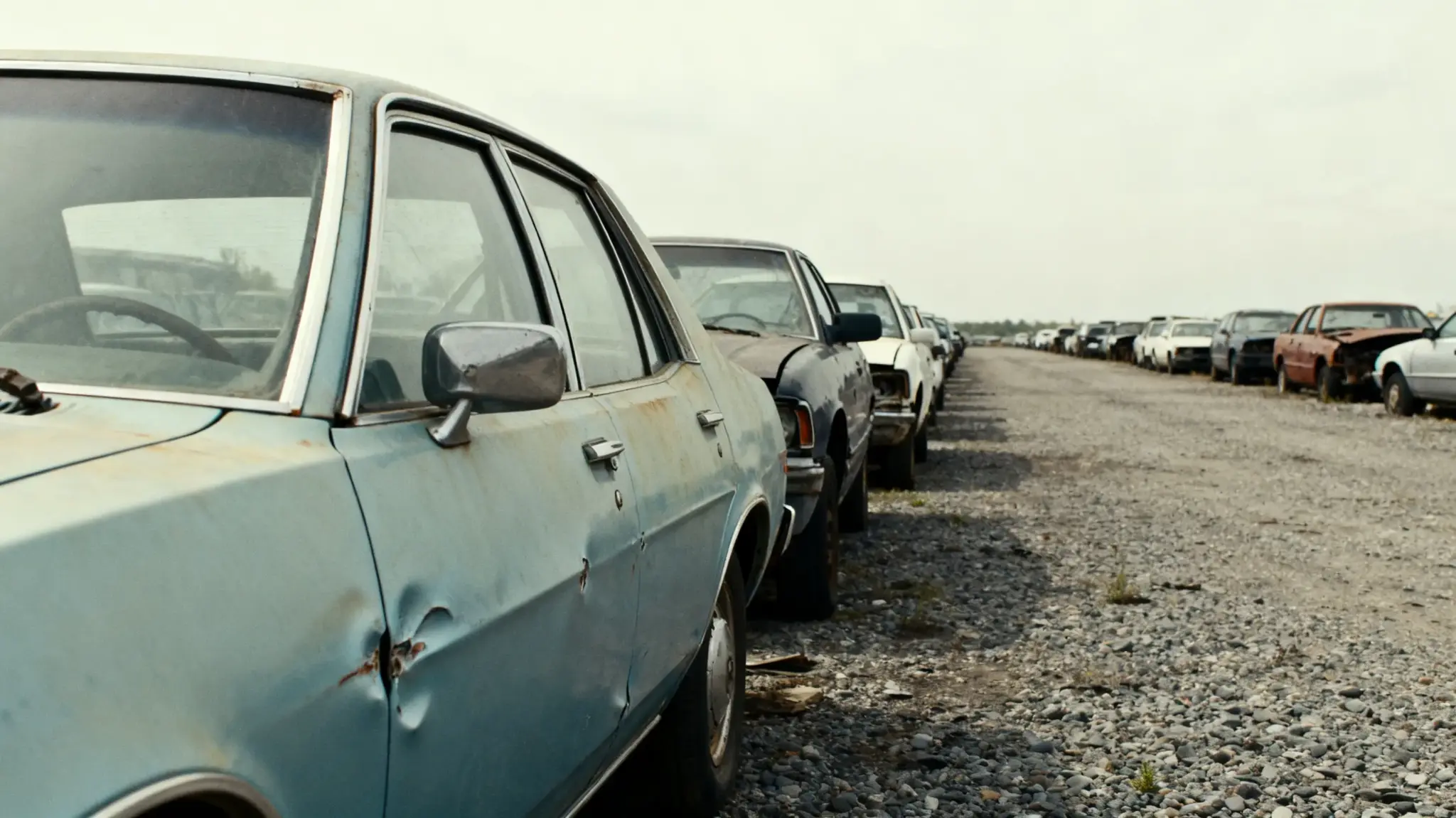 Row of rusted vintage cars parked on gravel lot under cloudy sky