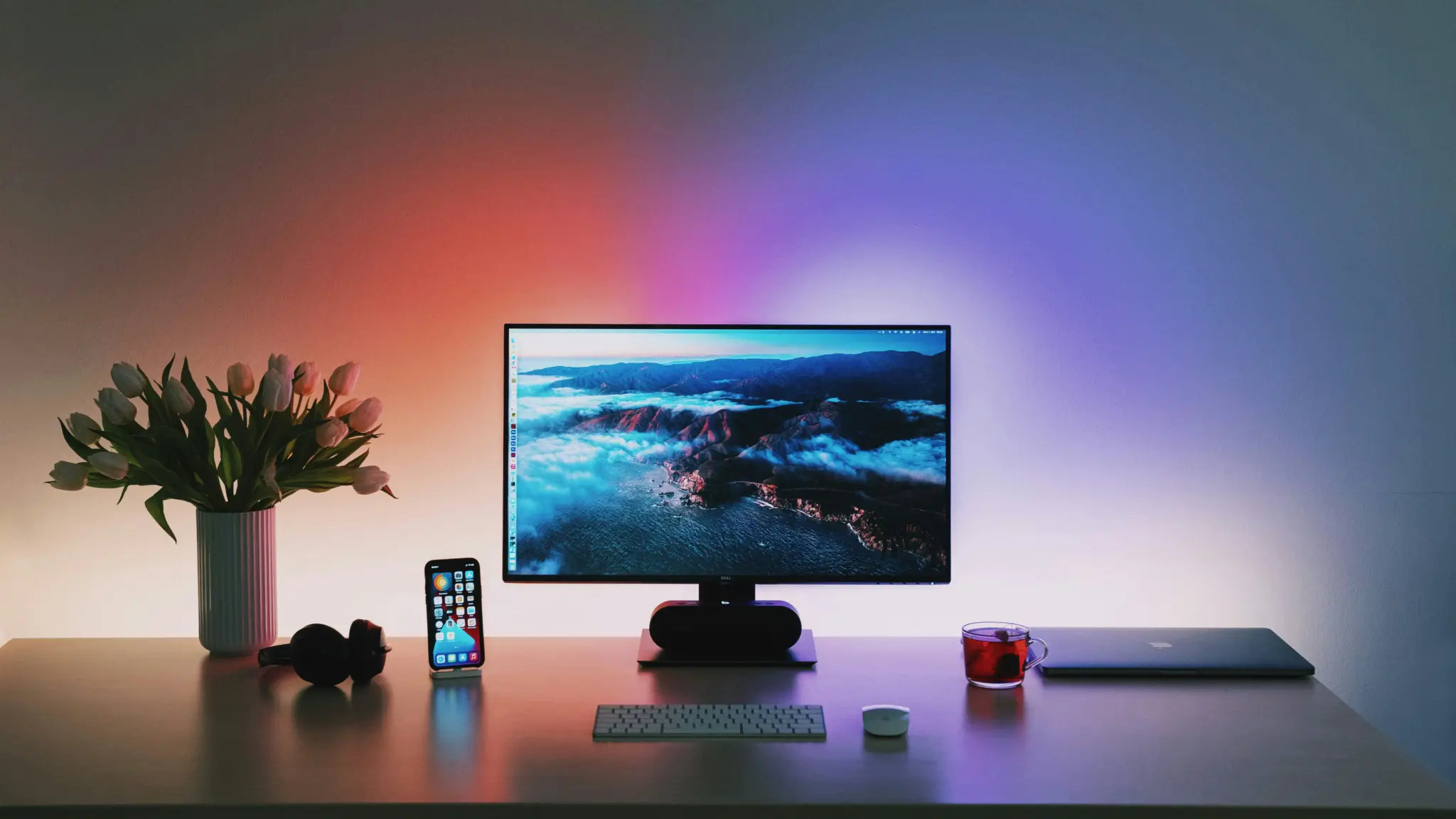 Computer monitor with landscape wallpaper on desk, surrounded by smartphone, headphones, and flowers in moody lighting