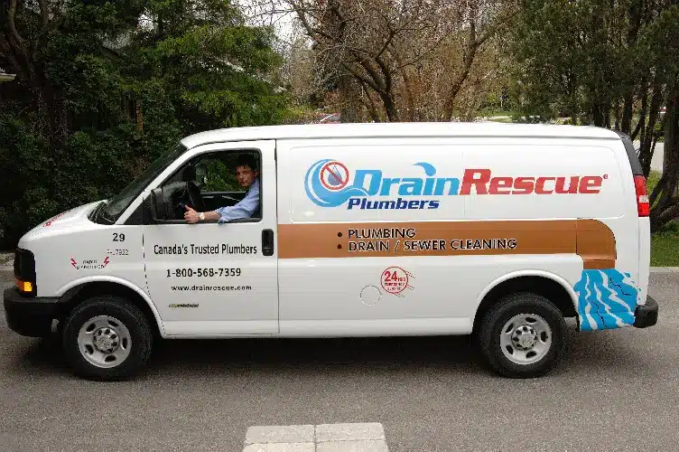 White plumbing service van parked on residential street, technician in driver's seat, trees in background