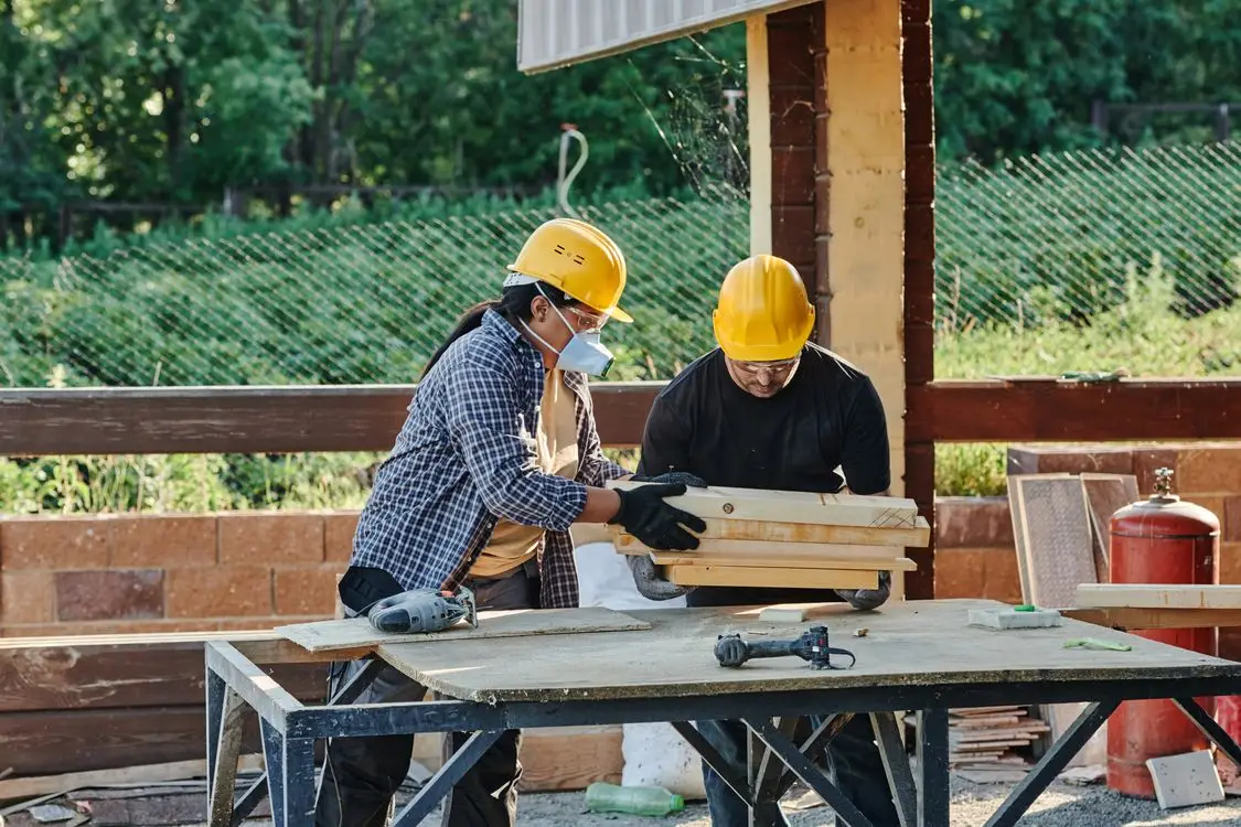 Construction workers wearing hard hats handling wooden planks at an outdoor worksite with power tools