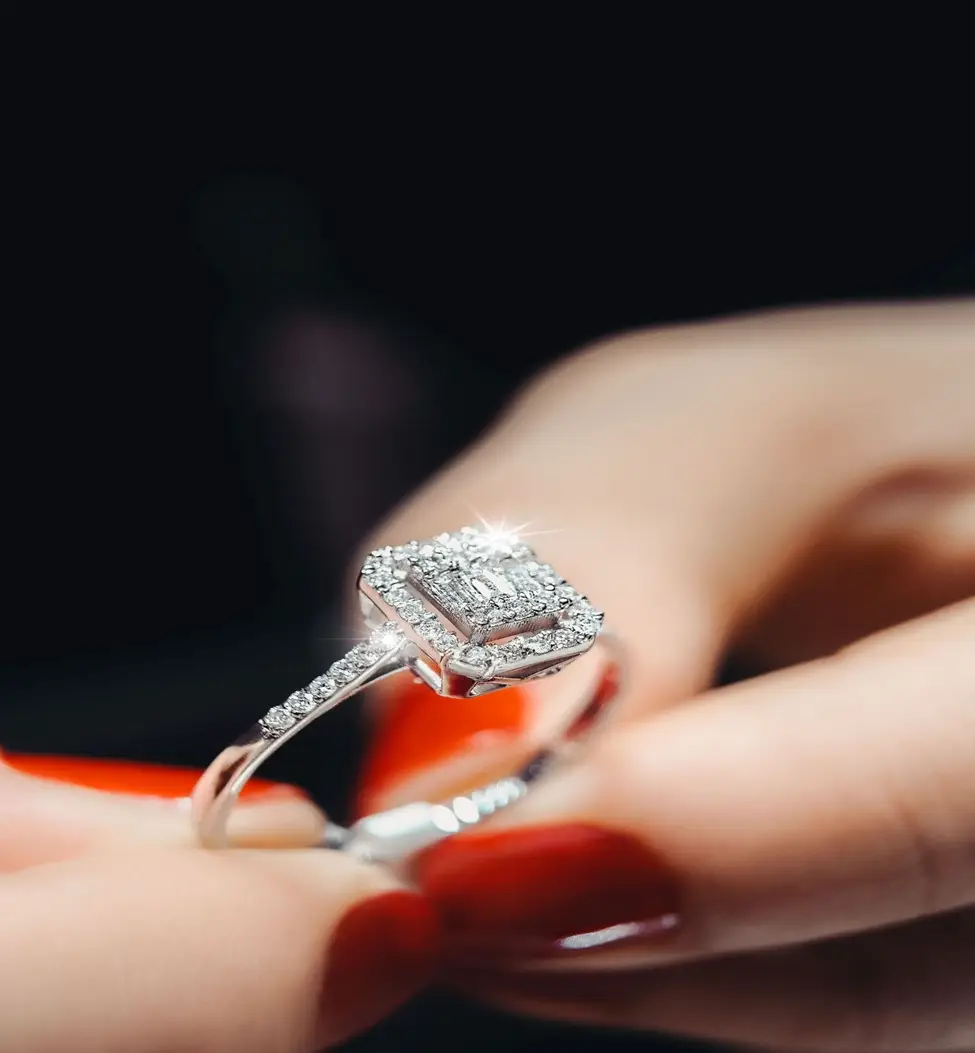 Square diamond ring with sparkling stones held between fingers against dark background