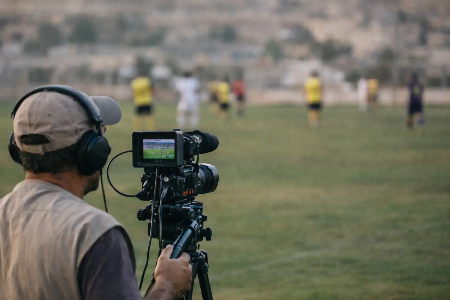 Camera operator filming outdoor soccer match on grassy field with blurred background players