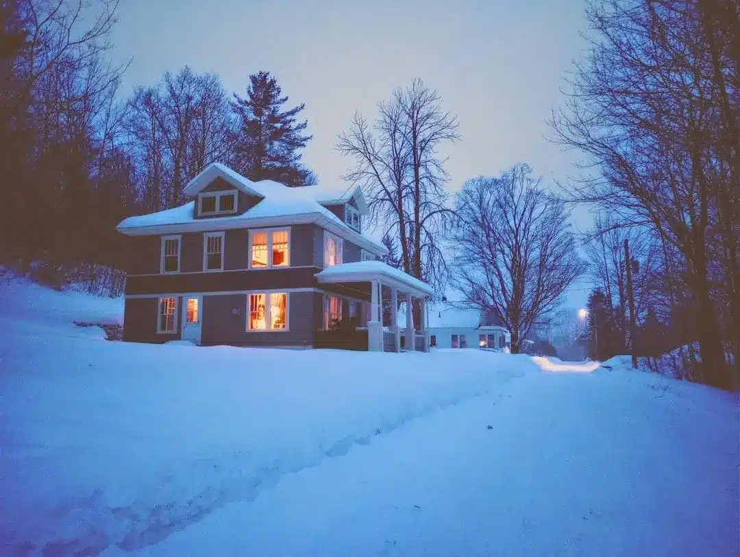 Large two-story house with glowing windows surrounded by snow-covered trees in twilight setting