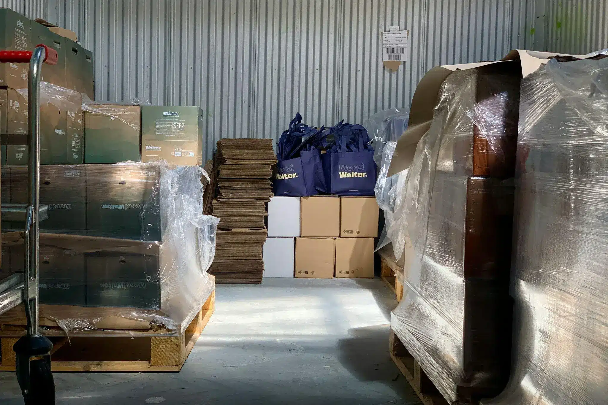 Stacked boxes, plastic-wrapped pallets, and folded cardboard in a sunlit warehouse space