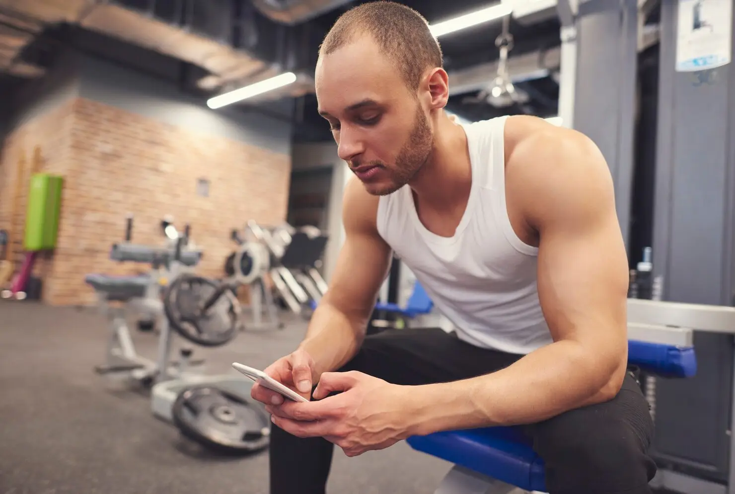 Man in white tank top using smartphone in gym with workout equipment in background