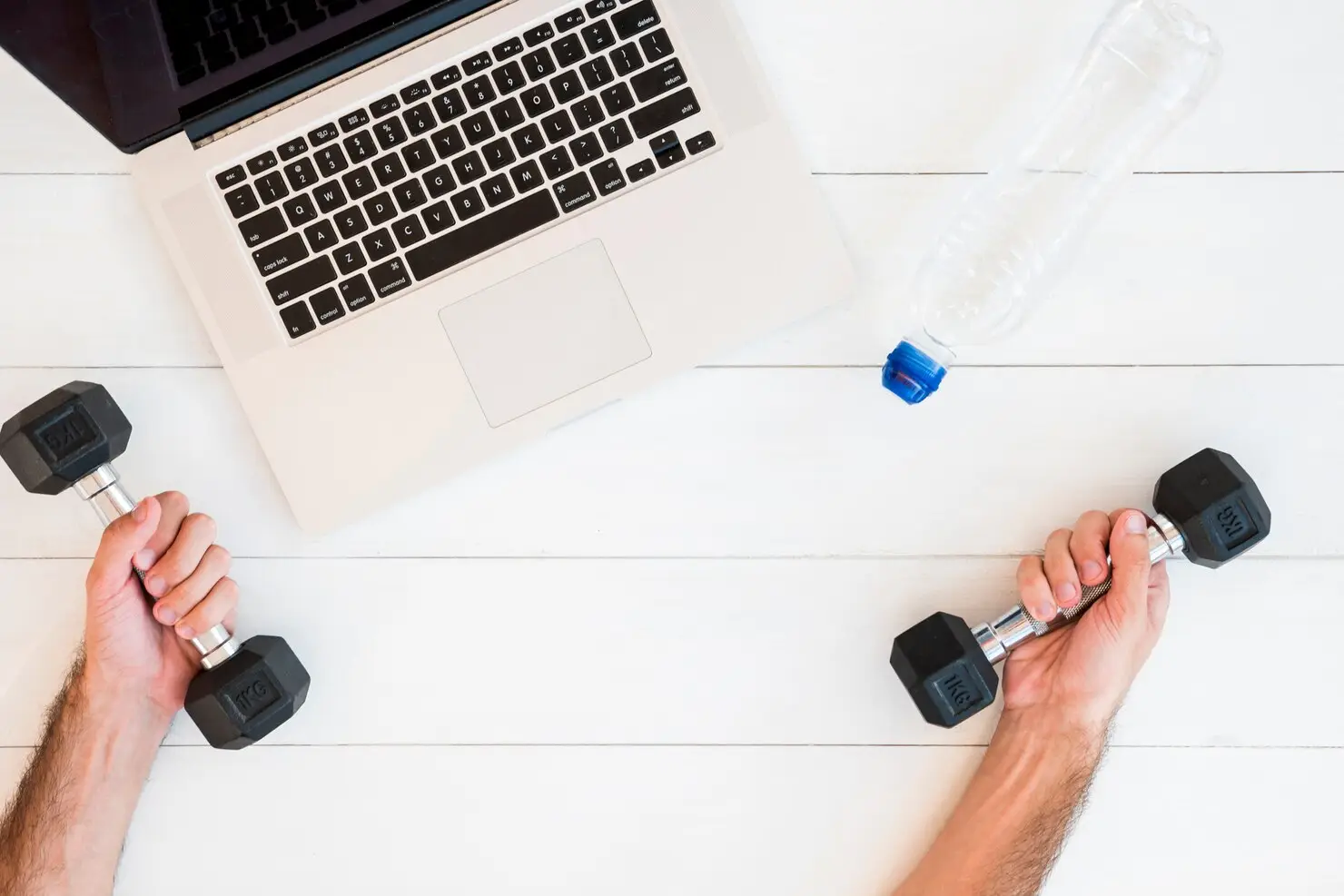 Laptop on white desk with hands holding dumbbells and water bottle nearby